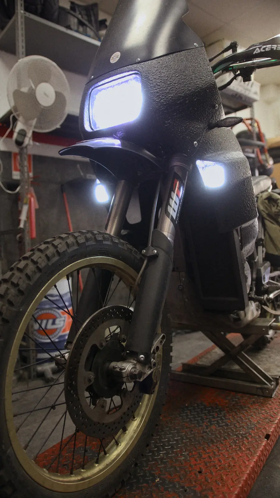 La moto nera di Bomber Garage sul ponte, coi fari accesi e la scritta oro sul serbatoio